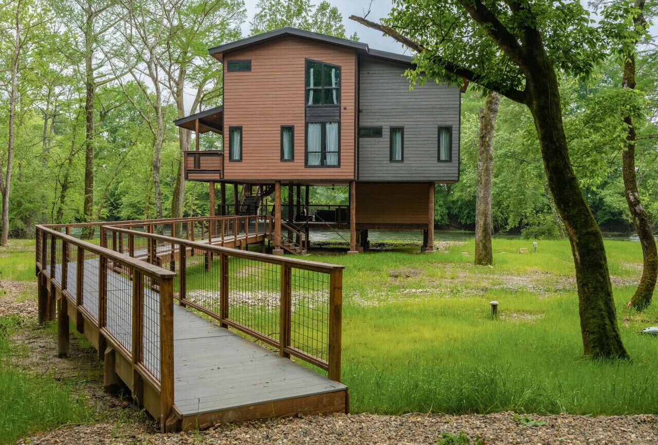 Une cabane en bois à deux étages, dotée d’une terrasse et d’un espace couvert, entourée d’une zone boisée.