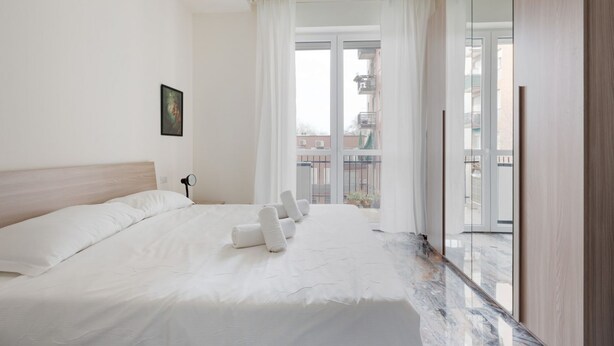 Habitación de hotel con una cama grande, una mesita de noche, una ventana con cortinas y vistas a edificios.
