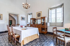 A dining room with a table set for four, surrounded by wooden chairs, a chandelier, and a large window with white shutters.