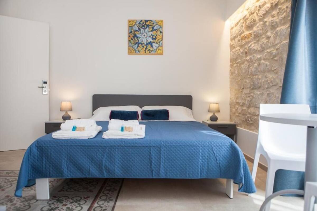 A neatly made bed with a blue bedspread, two pillows, and folded towels on the side. There is a stone wall and a white door in the background.