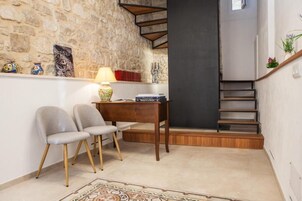 Een kamer met een stenen muur, een houten bureau met twee stoelen, een lamp en een wenteltrap.