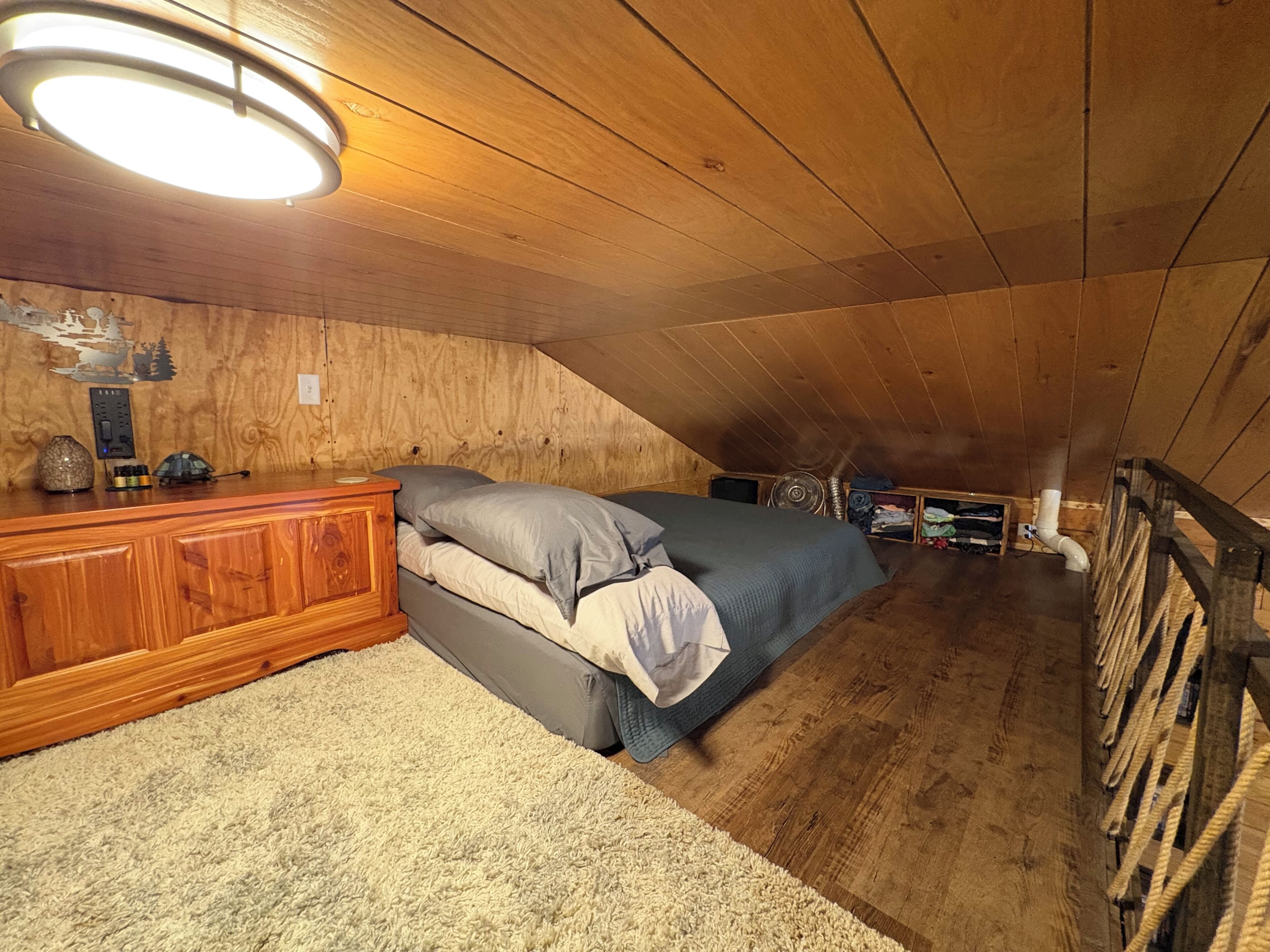 Un dormitorio tipo loft con techo inclinado, paredes de madera, cama con ropa de cama gris, cómoda de madera y una lámpara en el techo.