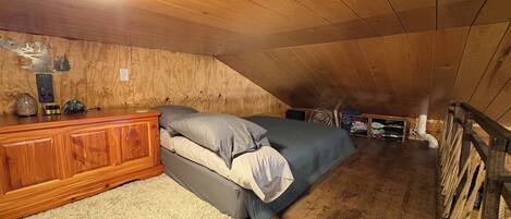 Un dormitorio tipo loft con techo inclinado, paredes de madera, cama con ropa de cama gris, cómoda de madera y una lámpara en el techo.