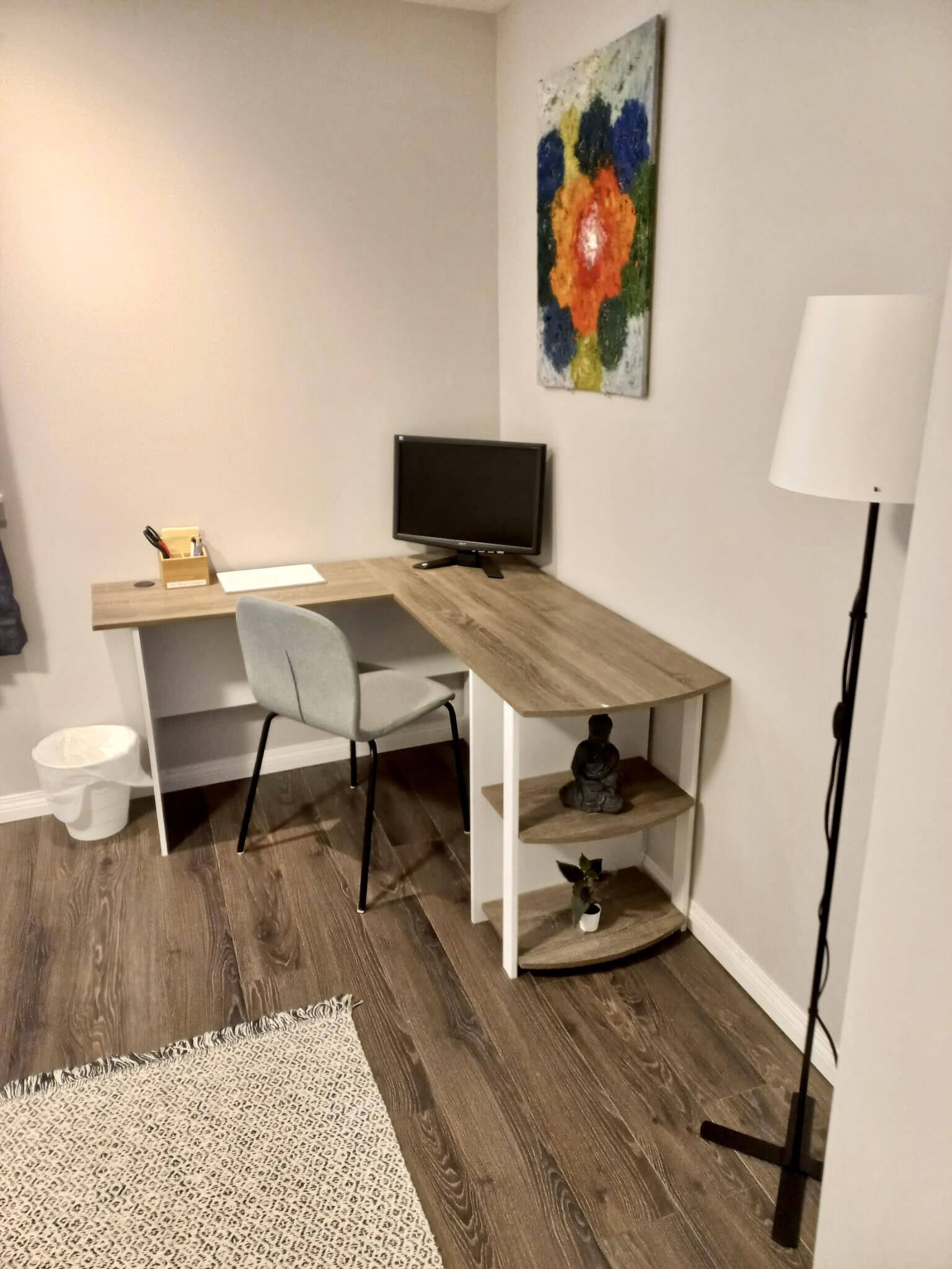 A modern office space with a wooden desk, a grey office chair, a computer monitor, a small statue, and a floor lamp.
