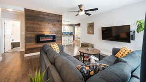 A modern living room with a fireplace, a ceiling fan, a flat-screen TV, and a dining area in the background. - Spacious downtown character rancher (Chilliwack)