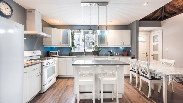 A modern kitchen with white cabinetry, a central island, and a dining area with white chairs. - Spacious downtown character rancher (Chilliwack)