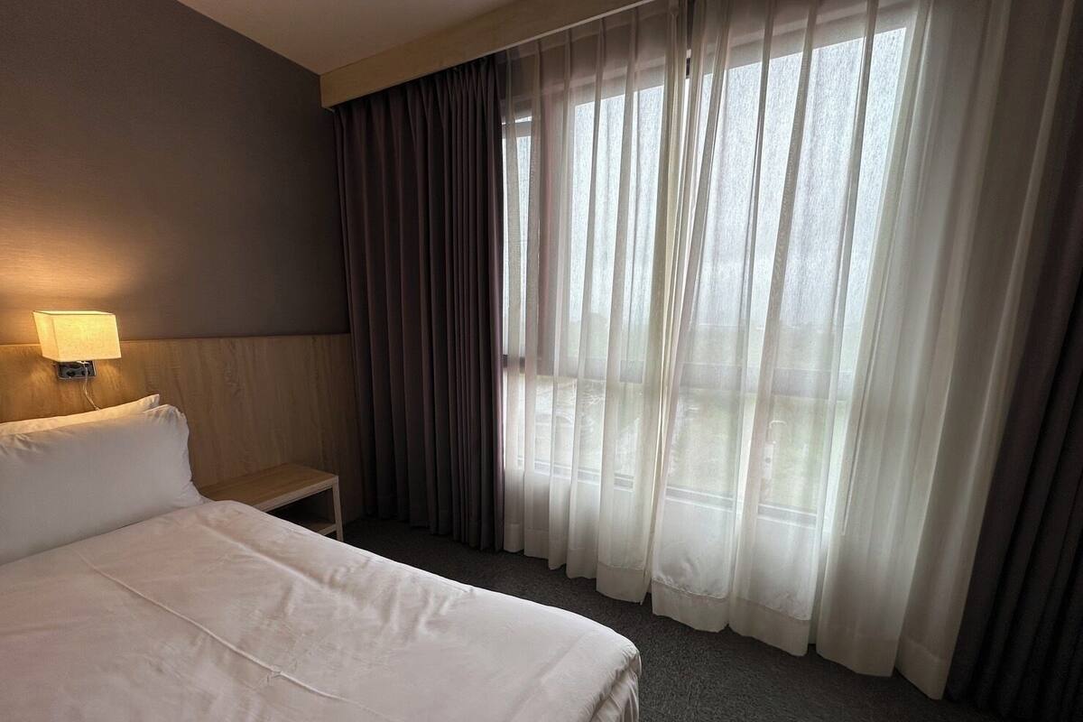 Hotellihuone, jossa on suuri sÀnky, yöpöytÀ, lamppu ja nÀkymÀ ohuiden verhojen lÀpi.