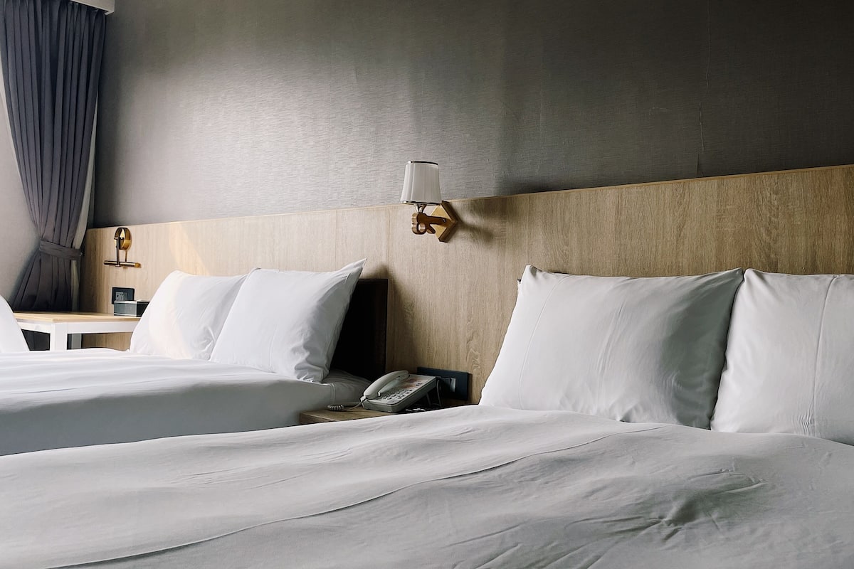Hotellihuone, jossa on kaksi sÀnkyÀ, puinen pÀÀty, yöpöytÀ lampun kanssa ja puhelin.