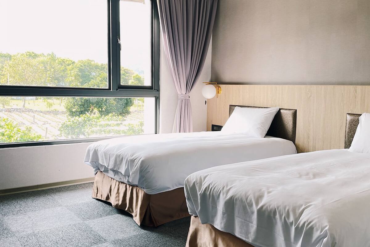 Hotellihuone, jossa on kaksi sÀnkyÀ, suuri ikkuna ja nÀkymÀ vehreÀÀn maisemaan.