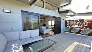 En udendørs terrasse med sofa, et glasbord og udsigt til huset.