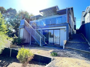 Une maison moderne à deux étages, dotée d’une façade en verre, d’un escalier en bois et d’une allée gravillonnée.