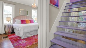 Une chambre avec un lit, une table de chevet, une lampe, une fenêtre avec des rideaux, un tapis rose et un escalier avec des marches violettes.