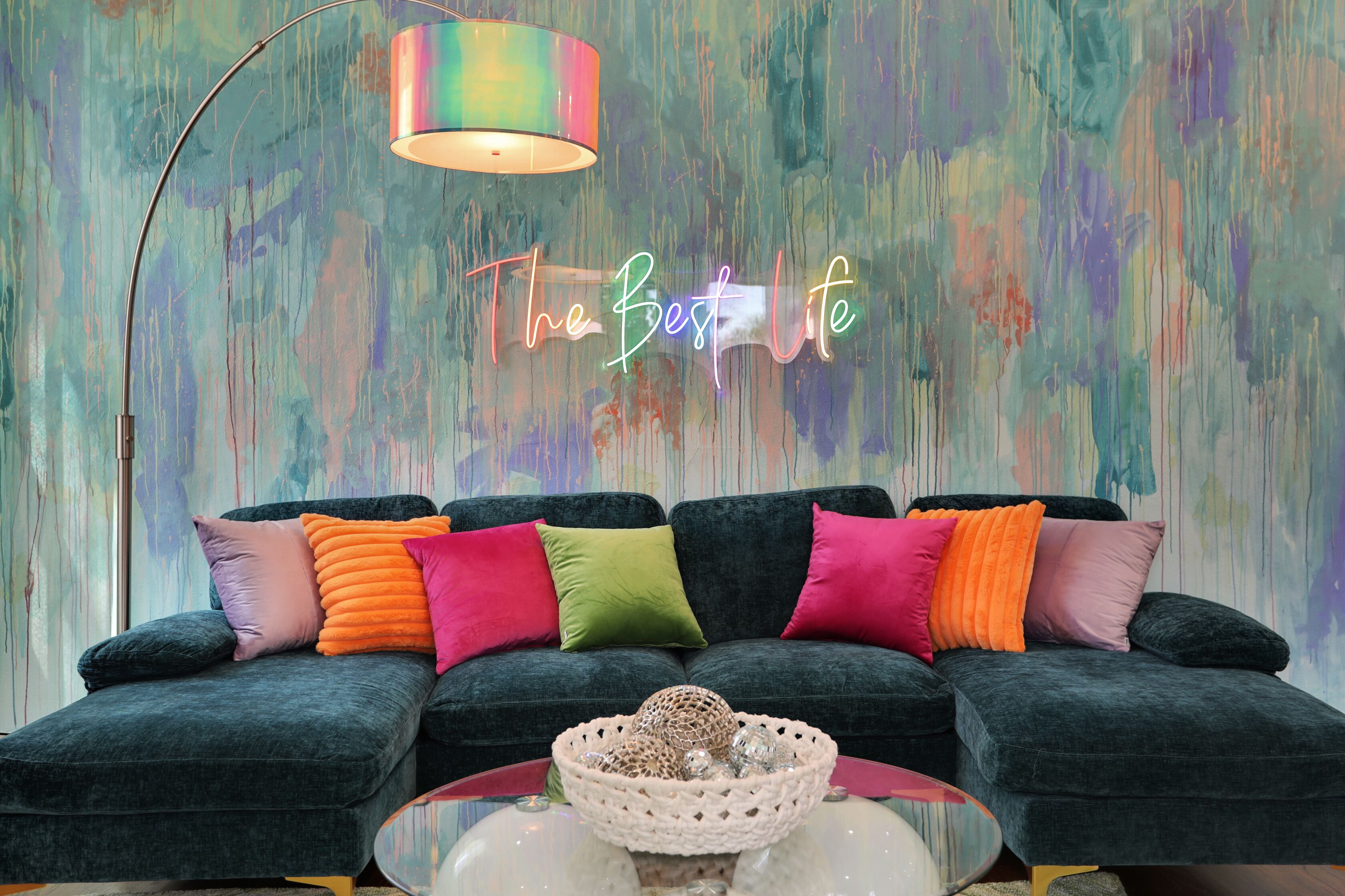 Un salon moderne avec un canapé vert foncé, des coussins colorés, une table basse en verre et un panneau lumineux au néon sur lequel on peut lire : « La meilleure vie qui soit ».