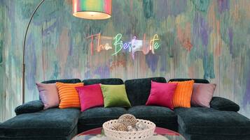 Un salon moderne avec un canapé vert foncé, des coussins colorés, une table basse en verre et un panneau lumineux au néon sur lequel on peut lire : « La meilleure vie qui soit ».