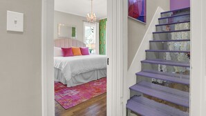 Une chambre à coucher avec un lit, des coussins colorés, un lustre et un escalier dont les marches sont violettes.