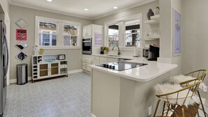 Une cuisine moderne avec des meubles blancs, un îlot central et un évier. On trouve des objets décoratifs sur les étagères et une poubelle près de l’entrée.