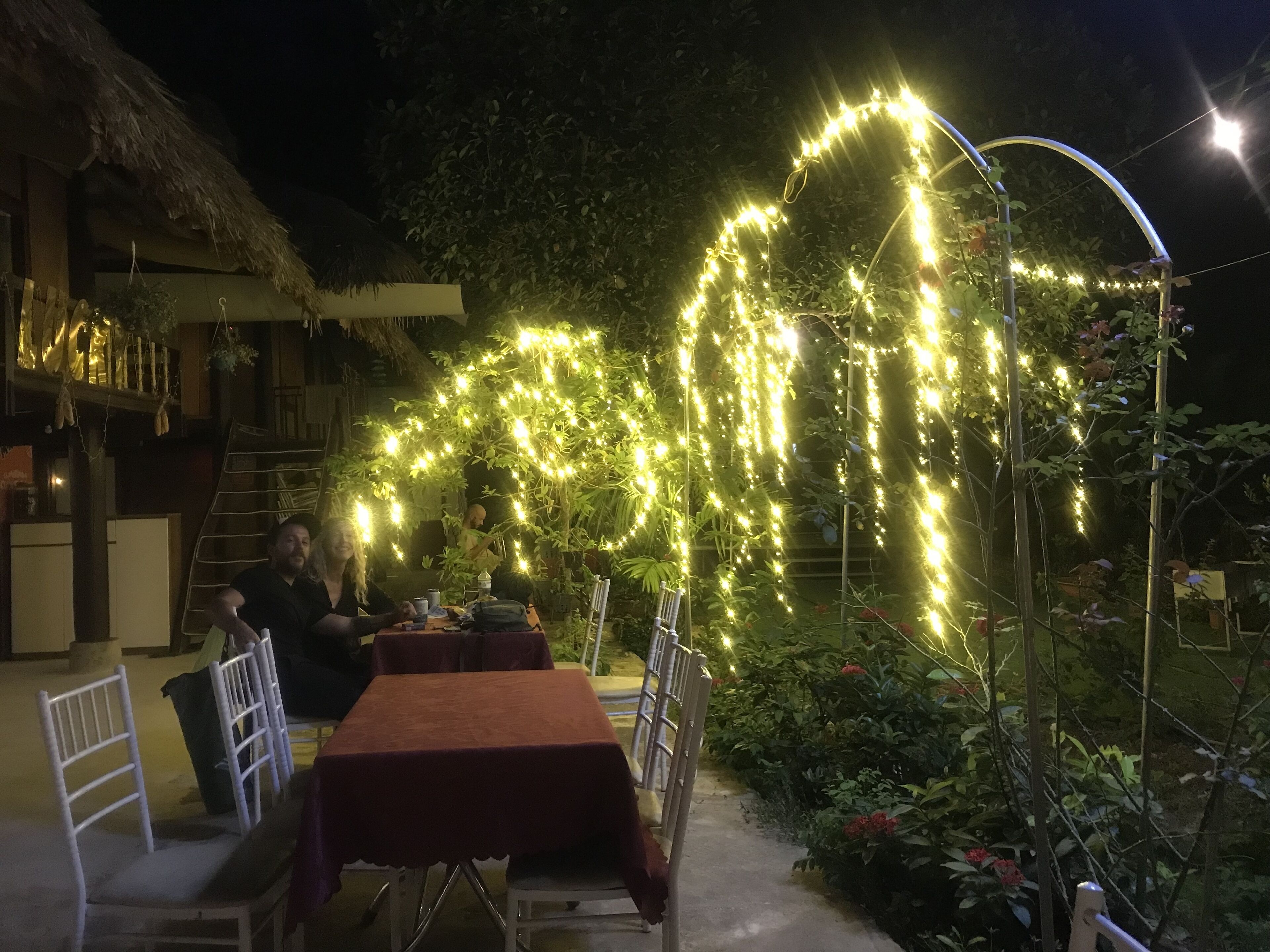 Un área de comedor al aire libre con una mesa y sillas, rodeada de follaje e iluminación con luces colgantes.