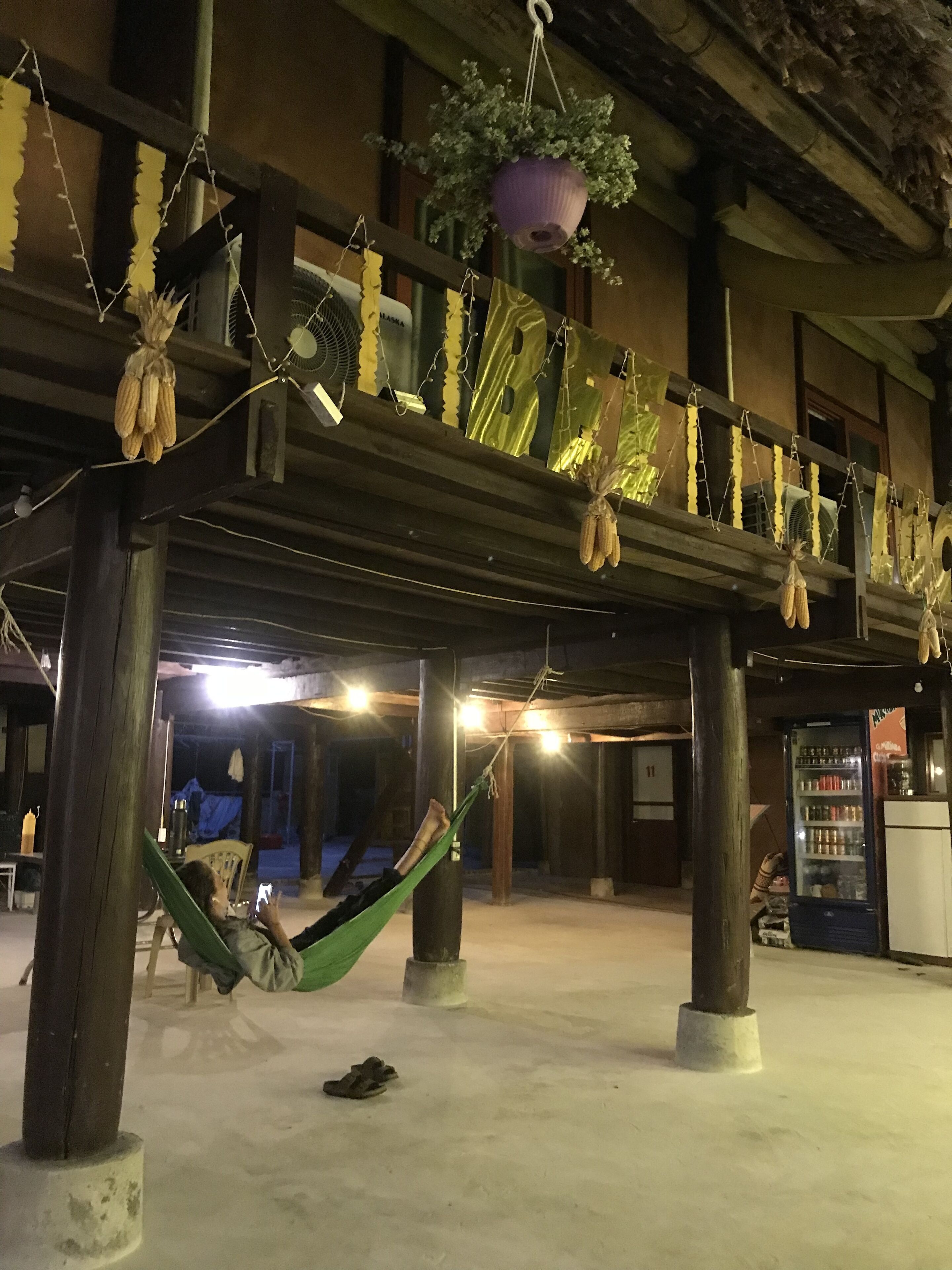 Une personne se détend dans un hamac, sous une structure en bois ornée de plantes suspendues et d’un panier suspendu.
