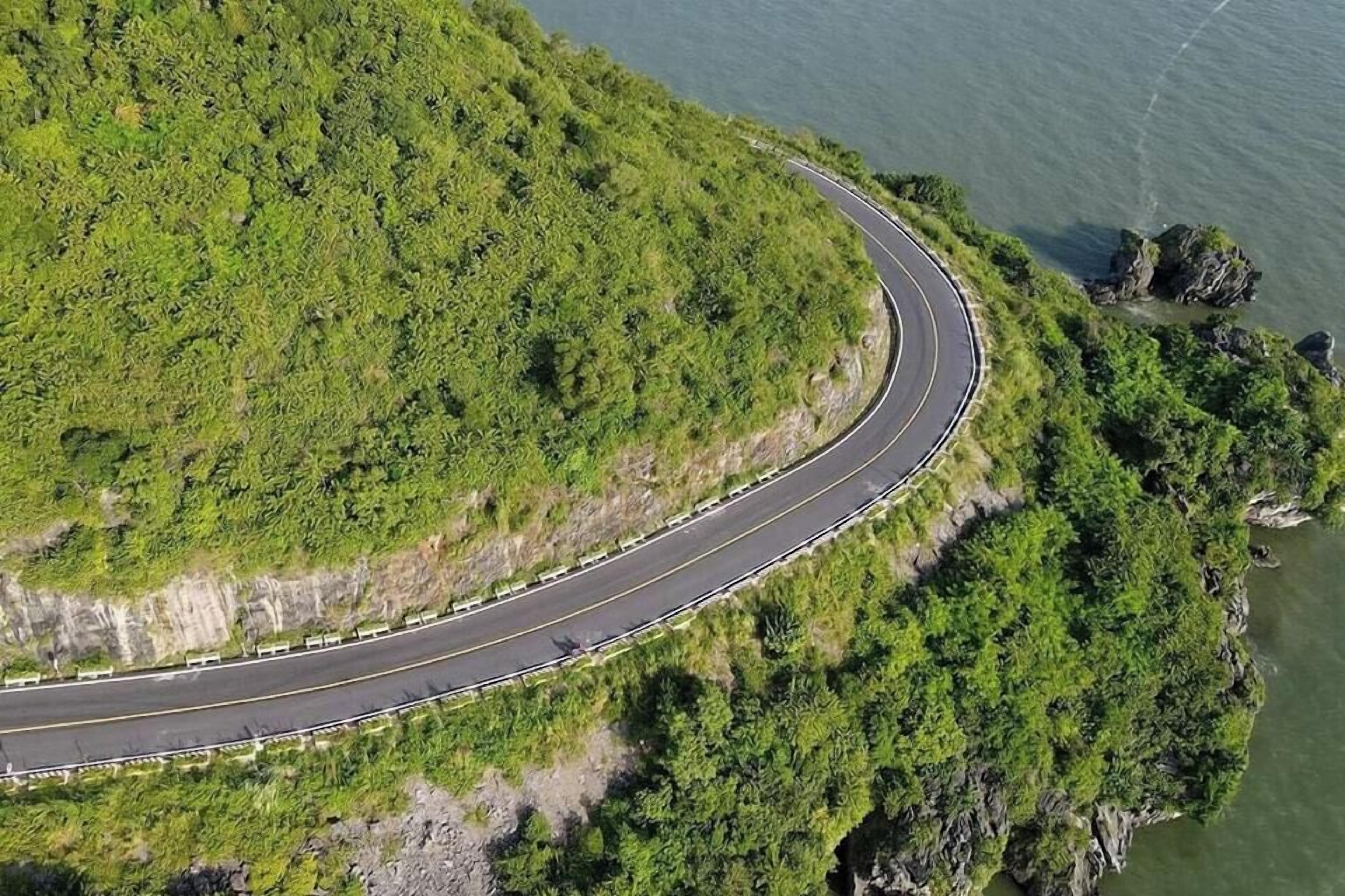 An aerial view of a winding road along a lush green hillside with a body of water on one side.
