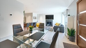 Uma sala de estar moderna com uma mesa de jantar de vidro, assentos confortáveis, lareira e área de cozinha ao fundo.