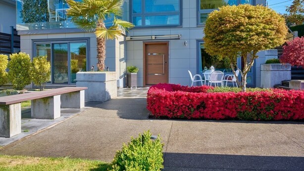 Uma casa moderna de dois andares com varanda frontal grande, porta de madeira e jardim bem cuidado com flores vermelhas e uma palmeira.