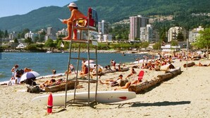 Uma praia com pessoas se banhando ao sol e nadando, uma torre de salva-vidas e edifícios ao fundo.