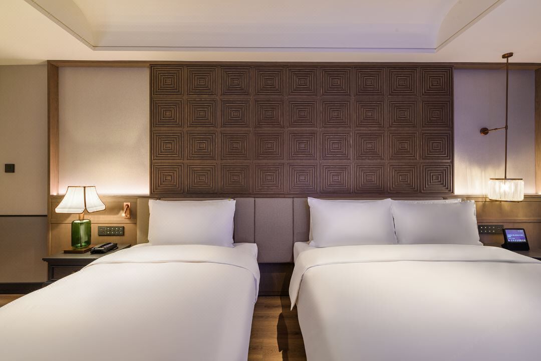 A hotel room with two beds, a wooden headboard, bedside lamps, and a nightstand with a phone.
