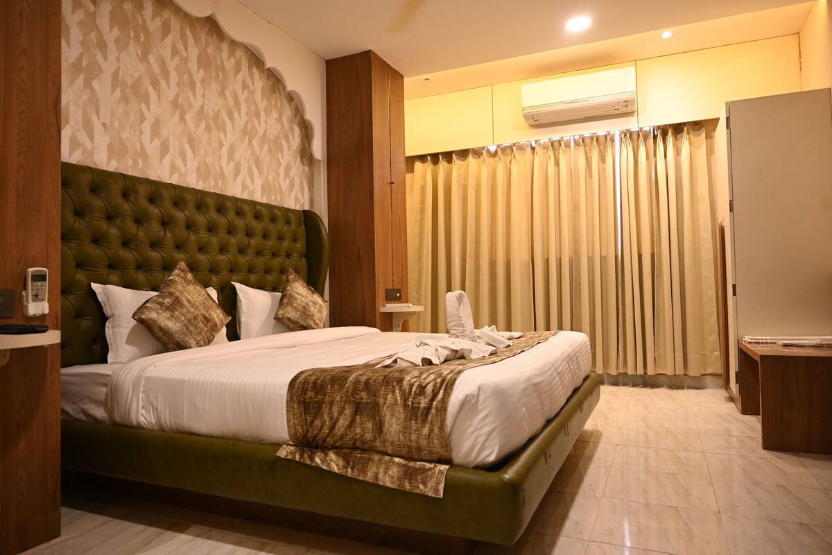A hotel room with a large bed, a wooden headboard, a built-in air conditioning unit, and a curtain-covered window.