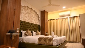 A hotel room with a green upholstered bed, a telephone on a wooden shelf, an air conditioning unit, and a curtain-covered window. - The Shah Imperial (Gwalior)