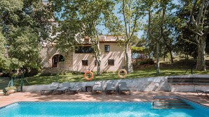 Une piscine avec des chaises longues, une bouée de sauvetage et un bâtiment en pierre, avec un jardin en arrière-plan.