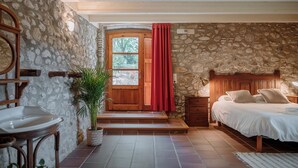 Une chambre confortable avec un mur en pierre, une porte en bois, un lit recouvert de draps blancs et une petite table avec une lampe.