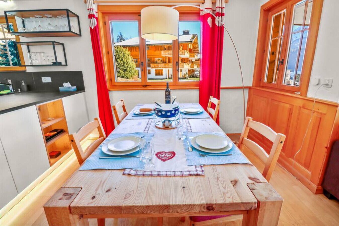 Un coin repas avec une table en bois dressée pour quatre personnes, une vue sur une montagne par la fenêtre et un espace cuisine avec des étagères et un luminaire suspendu.
