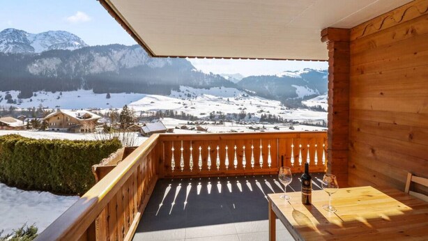 A wooden balcony with a view of snow-covered mountains and a village below. - Alexandra 6A - Spacious and comfortable accommodation (Château-d’Œx)