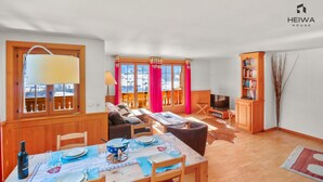 A modern living room with a dining area, wooden furniture, a bookshelf, and a view of a snowy landscape through large windows. - Alexandra 6A - Spacious and comfortable accommodation (Château-d’Œx)