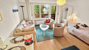 A cozy hotel room with a sofa, a small table with a bowl, a chair, a bed, and a window with a view of trees and a balcony.