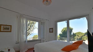 A bedroom with a large bed, a window with a view of trees and the sea, and a ceiling light. - Villa Salone 8 Emma Villas (Pollina)