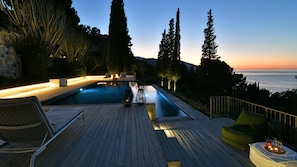 A serene outdoor pool area with a wooden deck, lounge chairs, and a view of the ocean at sunset. - Villa Salone 8 Emma Villas (Pollina)