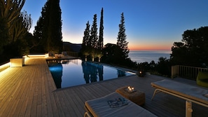 A serene poolside area with wooden decking, lounge chairs, and a view of the ocean at dusk. - Villa Salone 8 Emma Villas (Pollina)
