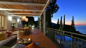 A terrace with a dining area, seating, and a view of the sea at dusk. - Villa Salone 8 Emma Villas (Pollina)