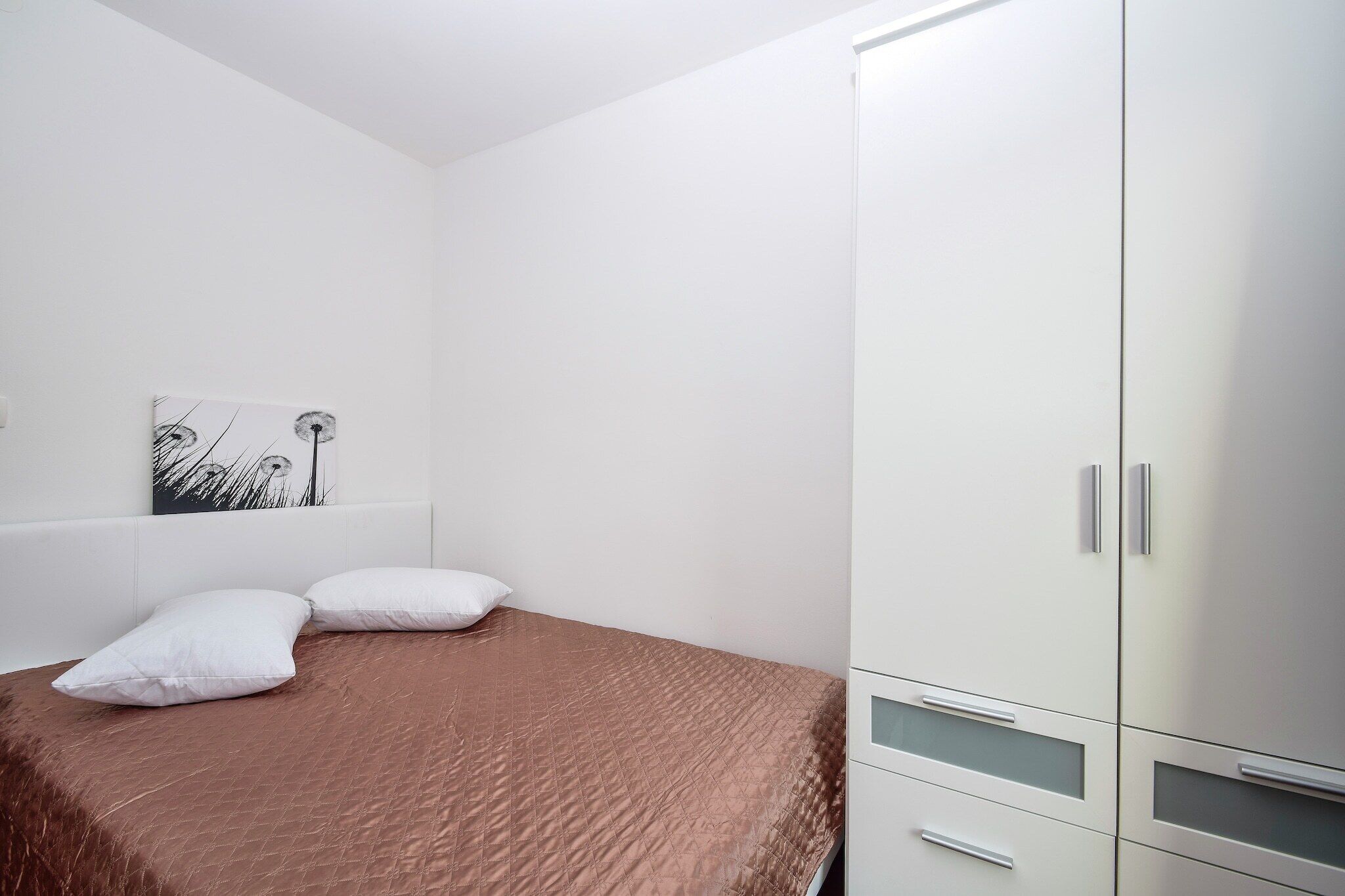 A bedroom with a bed, two pillows, a white wardrobe, and a wall-mounted artwork.