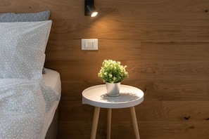 En seng med mønstret sengetøj, et lille hvidt bord med en potteplante og en vægmonteret lampe.
