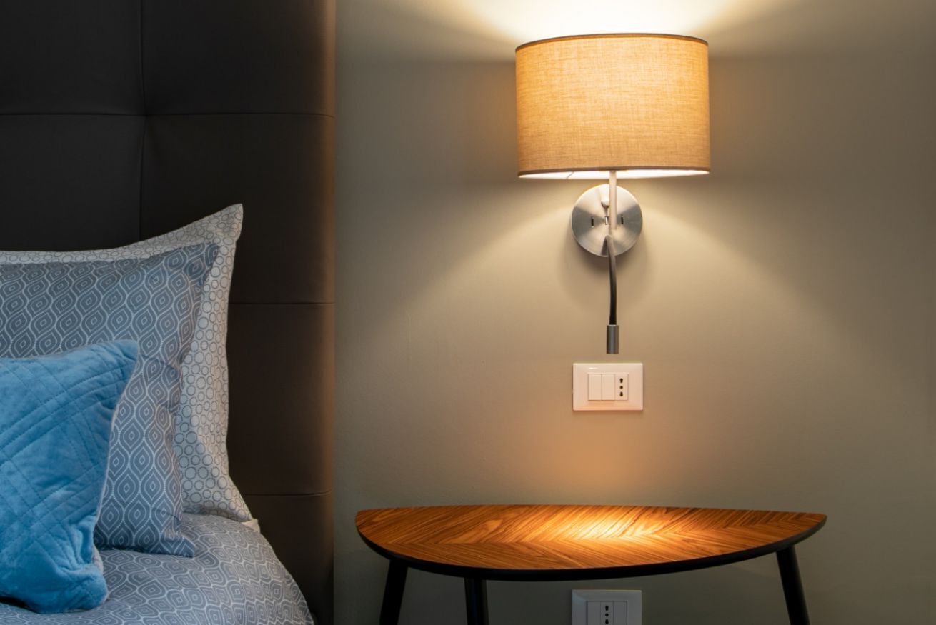 Una cama con almohadillas estampadas, una mesa redonda de madera y una lámpara fijada a la pared.