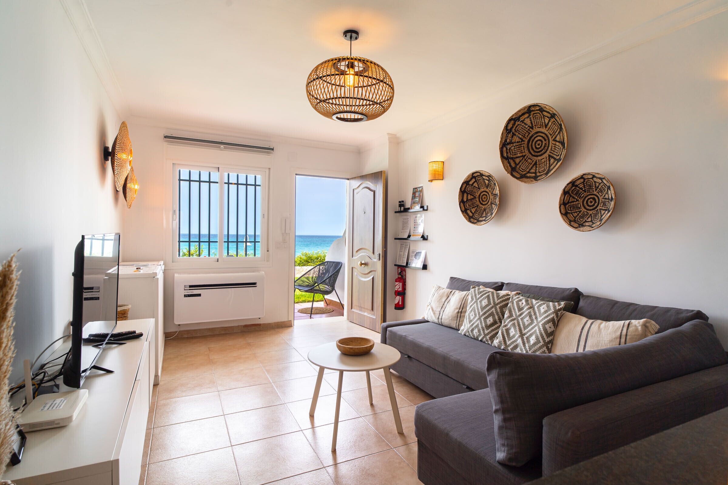 A living room with a sofa, a coffee table, a TV, and a view of the sea through the door.