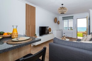 Eine moderne Küche mit integriertem Fernseher, eine Essecke mit grauem Sofa und Meerblick durch die offene Tür.
