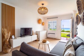 Een moderne woonkamer met een flatscreen televisie, een bank, een klein tafeltje en uitzicht op zee door een glazen deur.