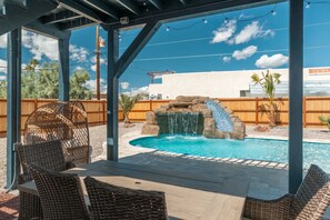 An outdoor pool area with a waterfall feature, wicker furniture, and a view of a white building in the background. - Epic Pool Retreat w/ Slide & Swim-In Grotto (Lake Havasu City)