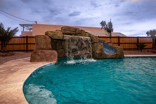 Una piscina con una cascada, rodeada por una valla de madera y palmeras.