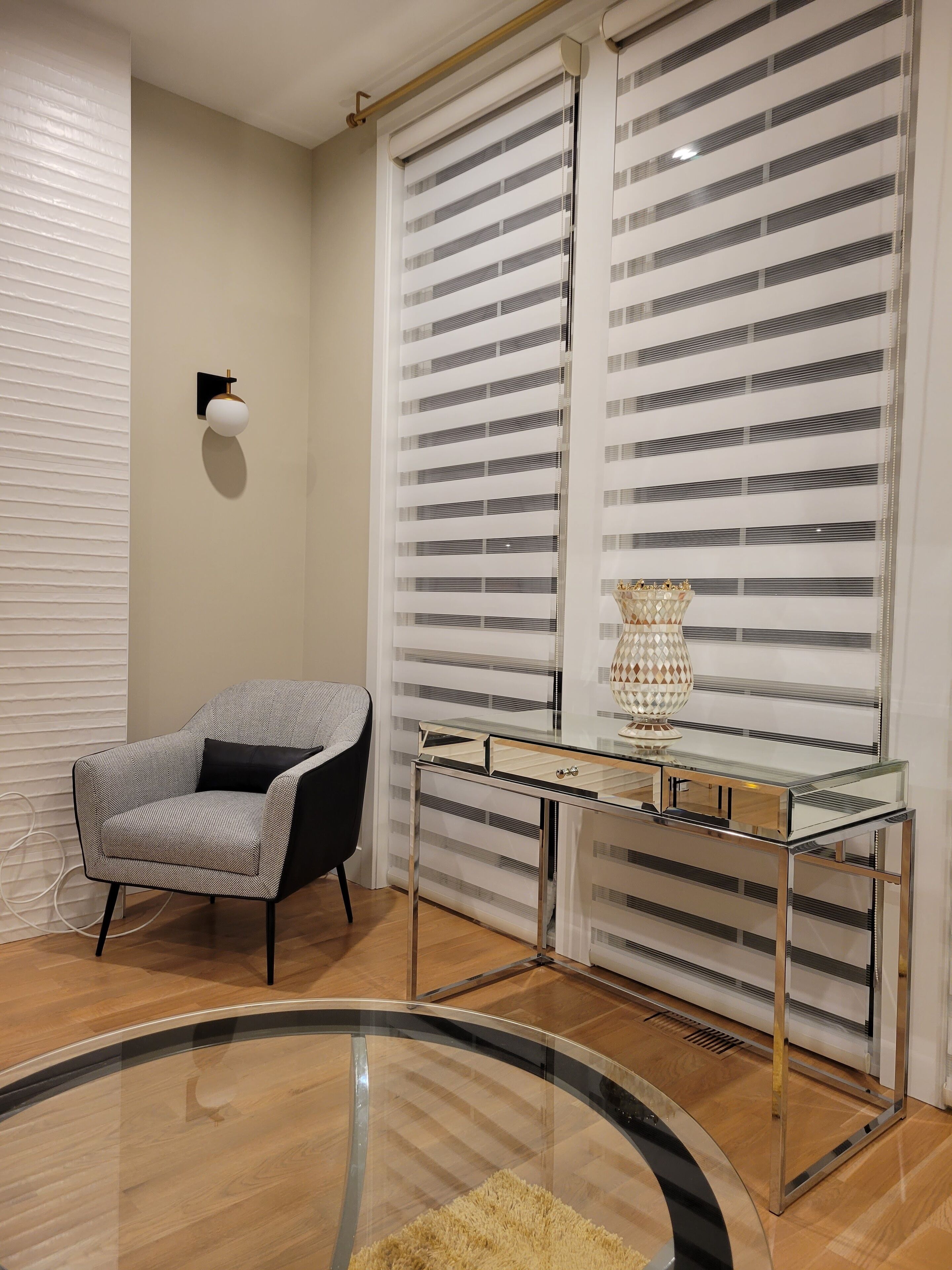 A modern room with a glass coffee table, a grey armchair, a glass console table with a decorative lamp, and striped blinds on the windows.