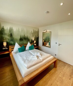 A bedroom with a large bed, a forest mural on the wall, a bedside table with a lamp, and a door.