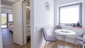 Quarto moderno e compacto com área de refeições, incluindo uma mesa redonda e duas cadeiras, relógio de parede embutido e janela com persianas.